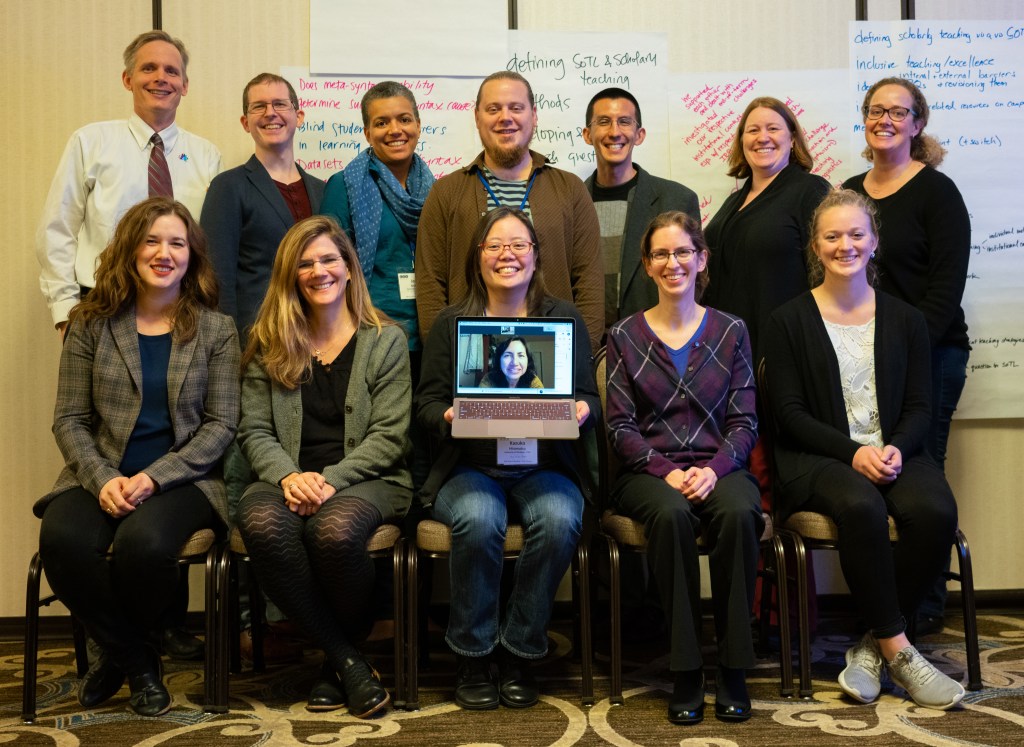 Group photo of twelve linguists in two rows. Seated in the front row, there are five women, and the middle one is holding a laptop with another linguist's face on it. The back row consists of seven people. 